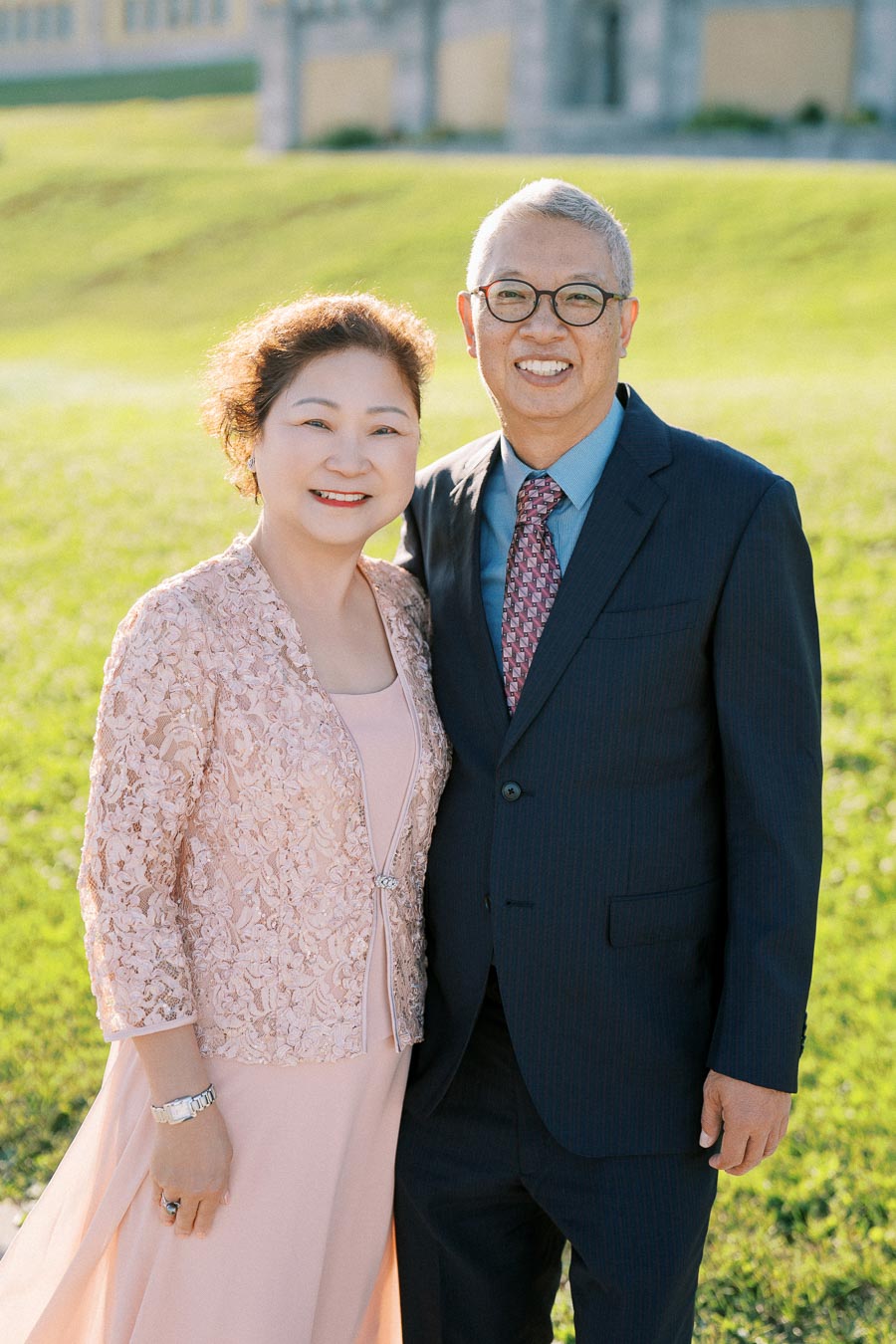 Smiling couple in formal attire posing outdoors on a sunny day with a grassy backdrop