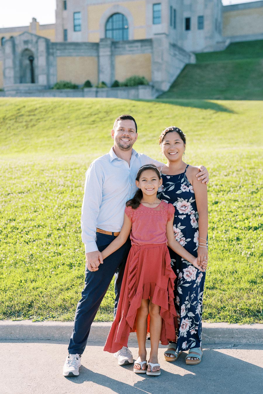 A happy family of three posing outdoors in front of a historic building, enjoying a sunny day.