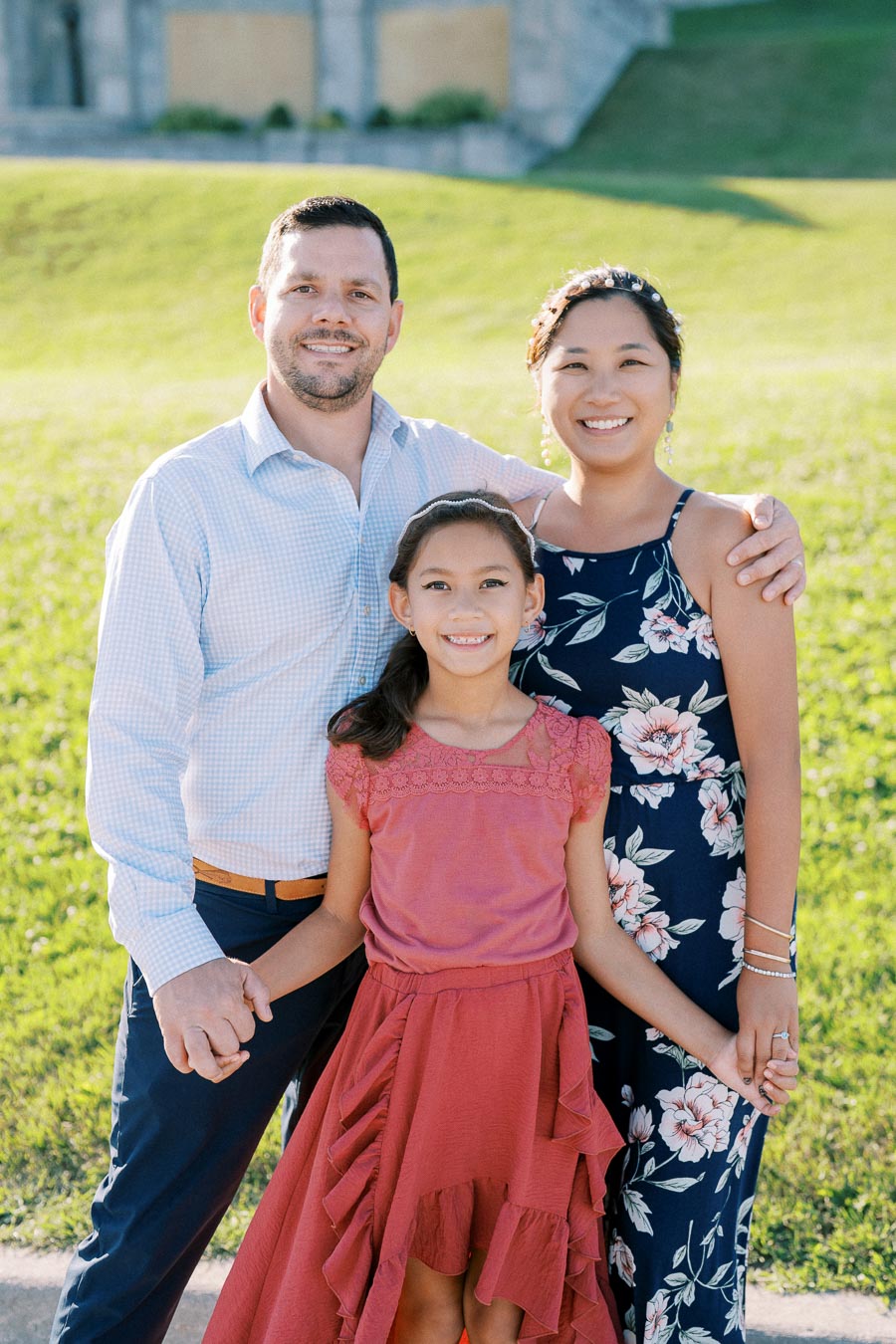 A happy family of three posing together outdoors on a sunny day, with a lush green background. The father wears a light blue shirt, the mother is in a floral dress, and the daughter is in a pink dress.