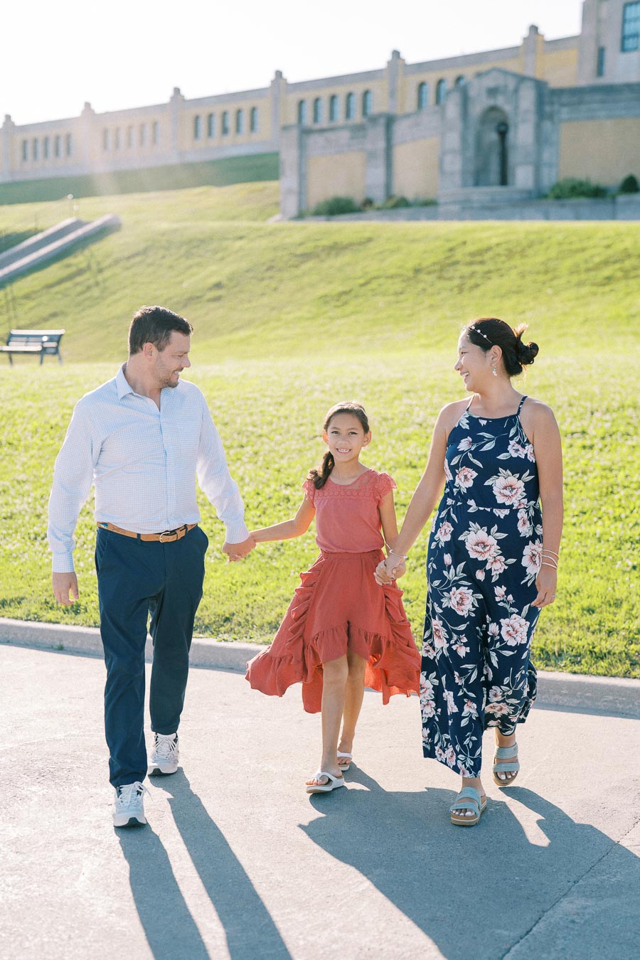 A family enjoying a sunny day at the park, walking hand in hand on a paved path with a historic building in the background.