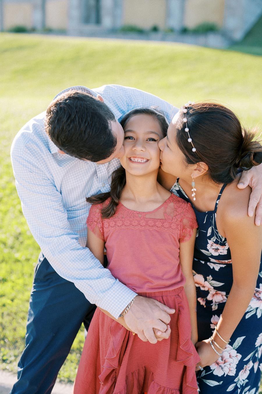 A family enjoying a sunny day in the park, with parents lovingly kissing their smiling daughter who is wearing a red dress and braces.