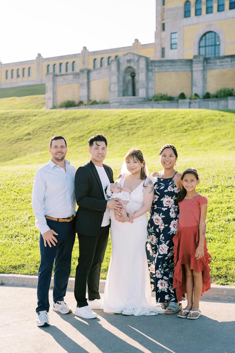 A diverse group of five people, including a couple holding a baby, posing happily in front of a historic building on a sunny day, showcasing family and togetherness in an outdoor setting.