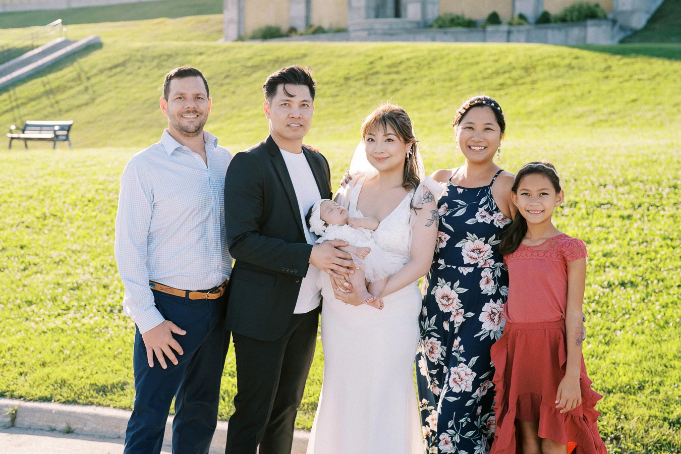 A group of six people, including a baby, posing outdoors in a park during a sunny day. One individual holds the baby, while others stand beside them, all smiling. The background includes green grass and trees, creating a joyful and family-oriented atmosphere.