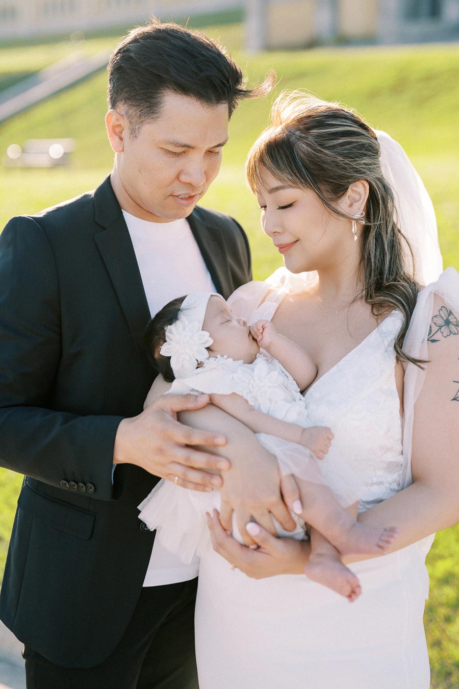A loving couple in formal attire holding their sleeping baby in a sunlit outdoor setting, symbolizing family unity and warmth.