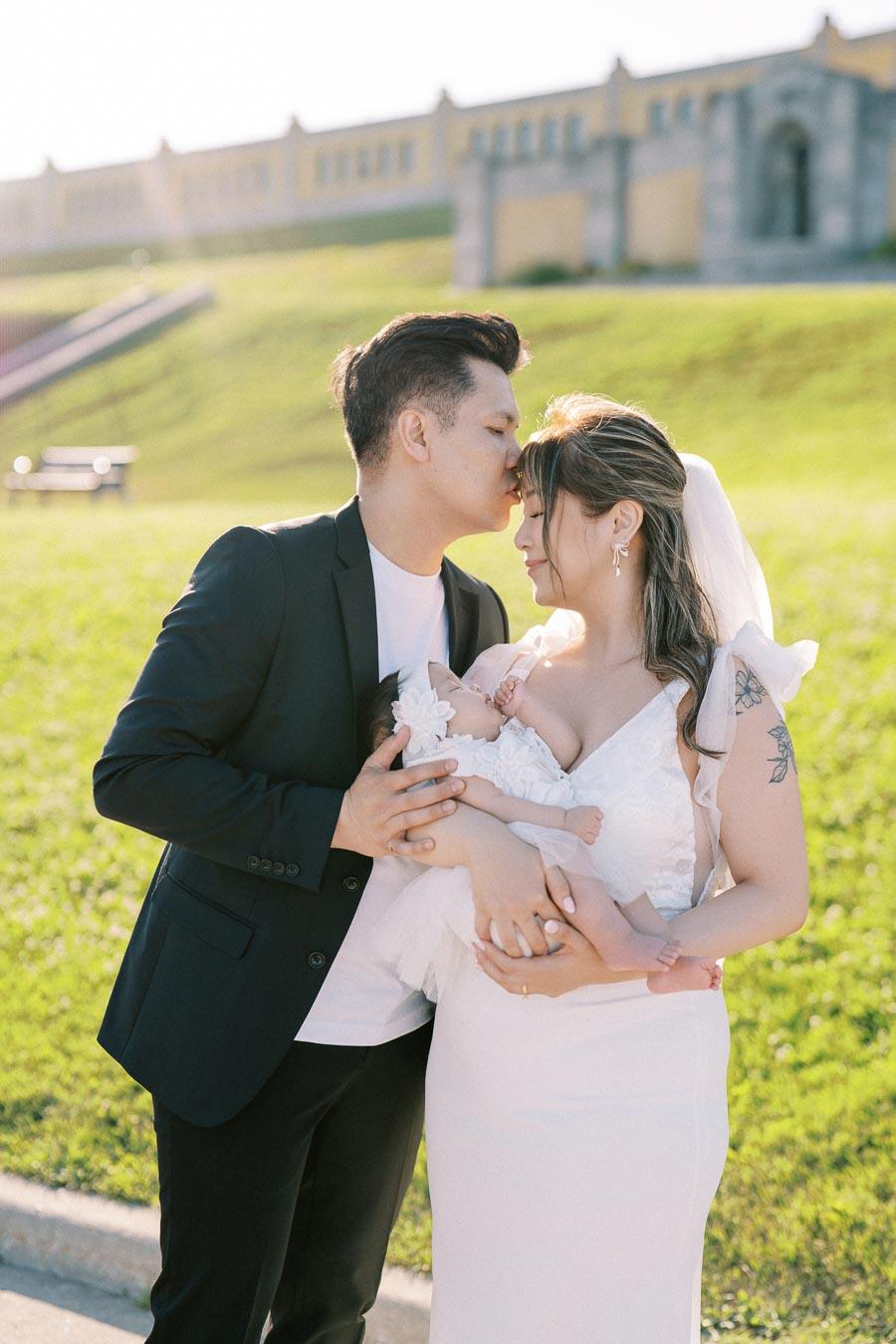 A loving couple embracing and holding their baby in a serene outdoor setting, with the man gently kissing the woman on the forehead. She is wearing a white dress and veil, while he is dressed in a dark suit. The sunlit background features green grass and historic architecture, creating a peaceful and joyful atmosphere. Perfect image for family and love-themed content.