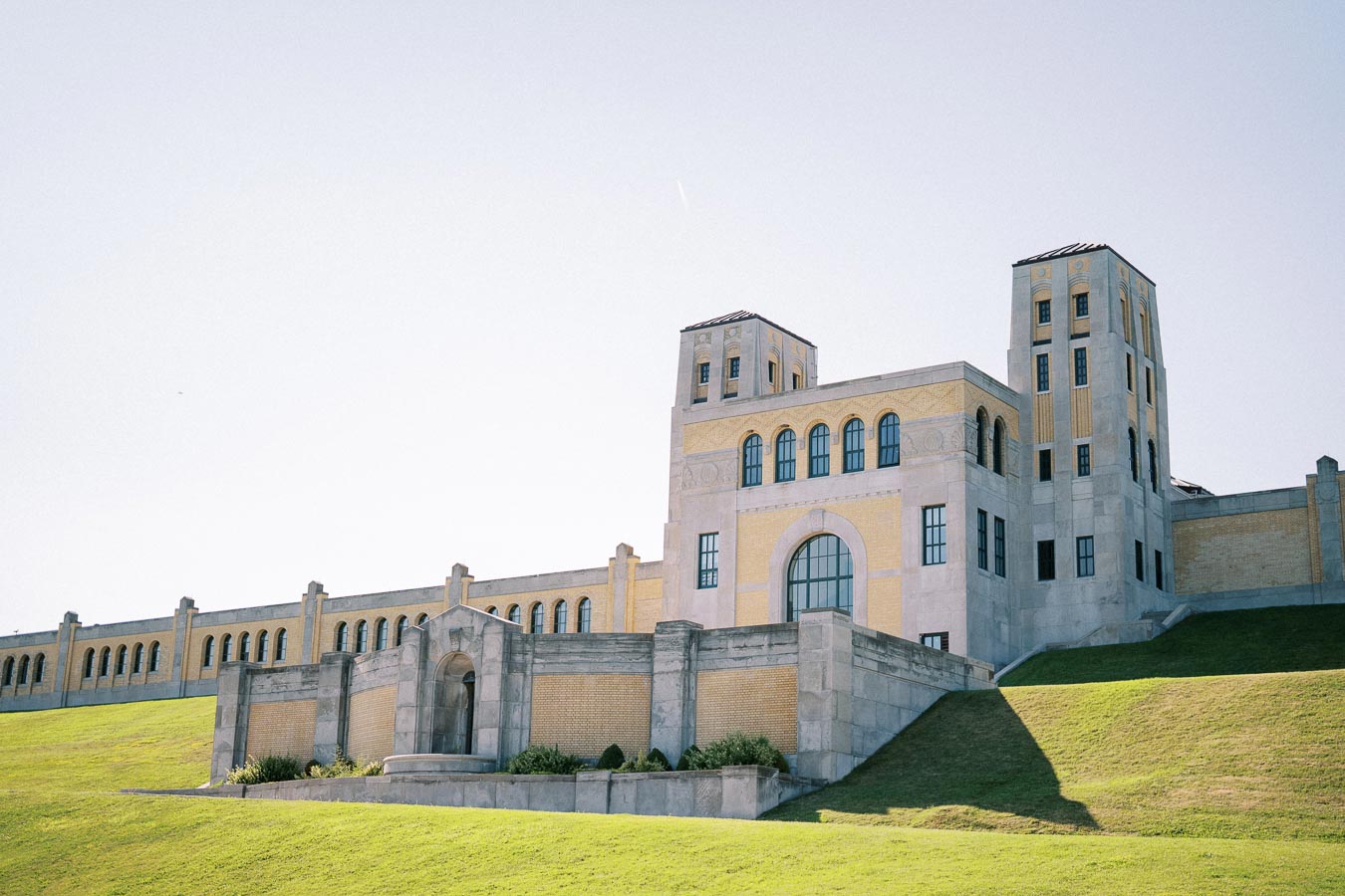 Historic architectural building with tall towers and arched windows set on a green hillside under a clear blue sky.