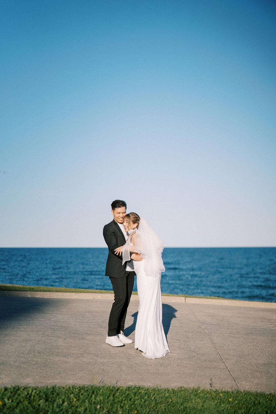 Elegant couple embracing on their wedding day by the ocean, under a clear blue sky, with the bride in a flowing white dress and veil, and the groom in a sleek black suit.