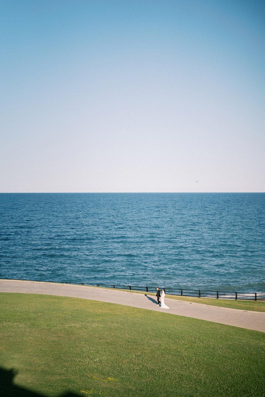 A bride and groom walking along a scenic oceanfront path with a clear blue sky and expansive sea in the background, capturing a serene and romantic moment.