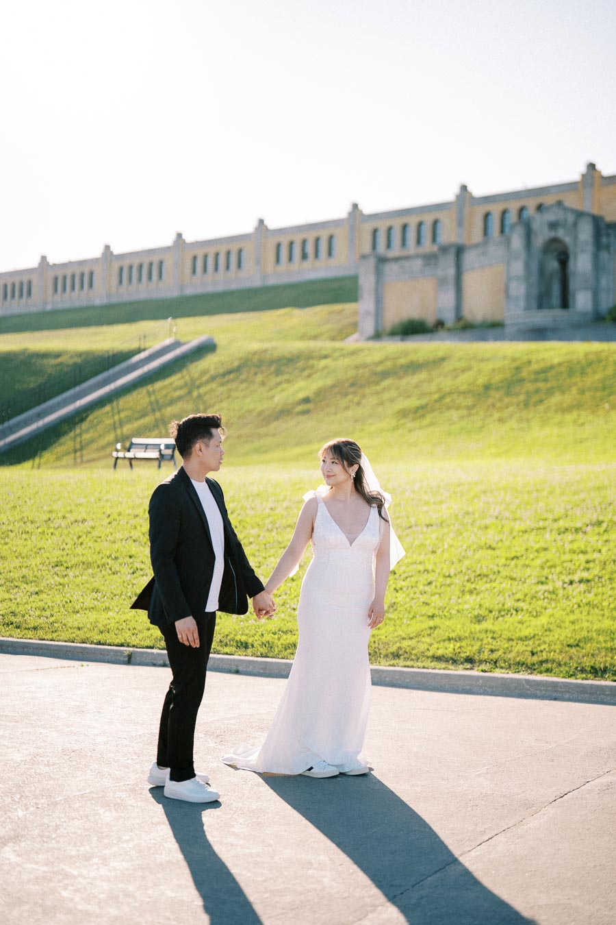 A bride and groom holding hands outdoors on a sunny day, with a grassy hill and historic building in the background.