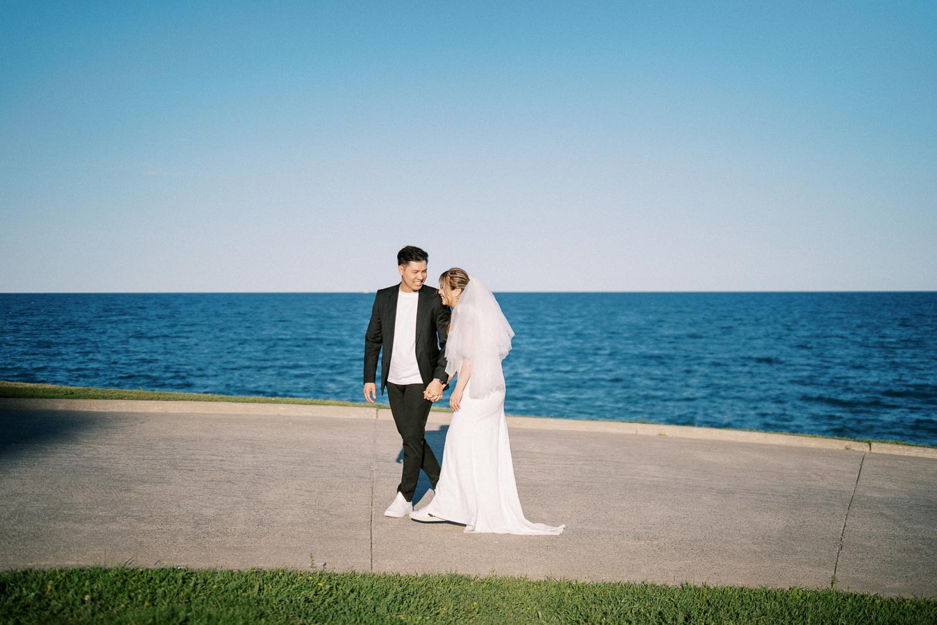 Bride and groom walking hand in hand by the ocean on a clear day.