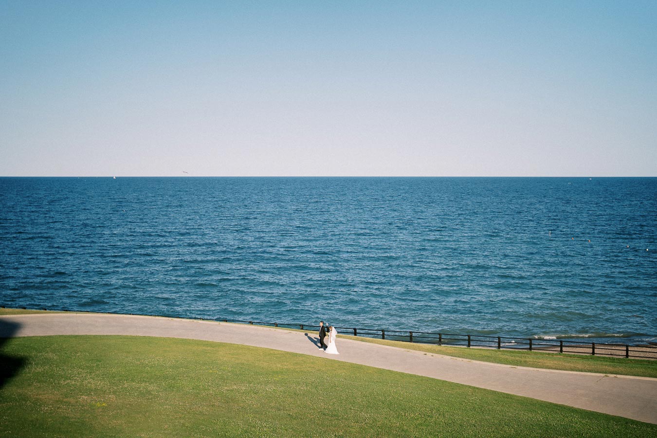 Bride and groom walking on a pathway by a serene blue ocean on a sunny day, surrounded by green grass and a clear sky.