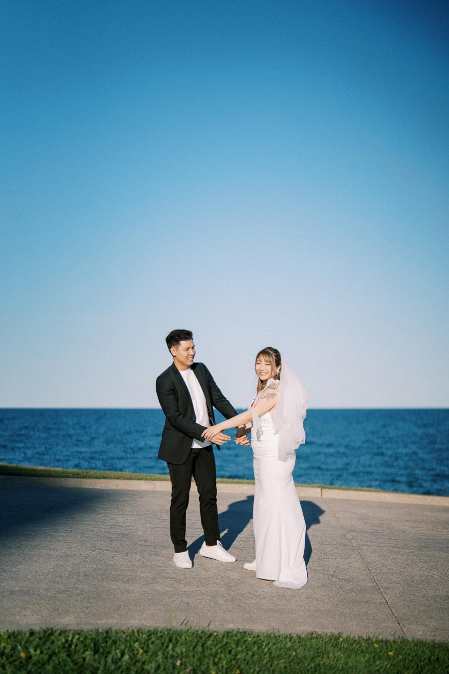 Bride and groom holding hands by the ocean, standing on a sunny day with clear blue skies.