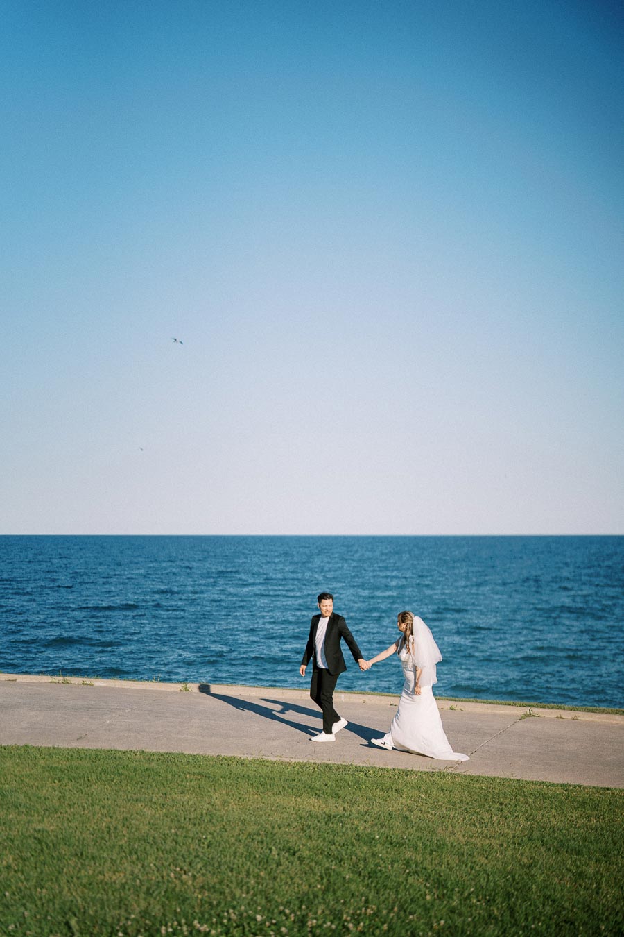 Bride and groom holding hands walking by the ocean on a clear sunny day with blue sky.