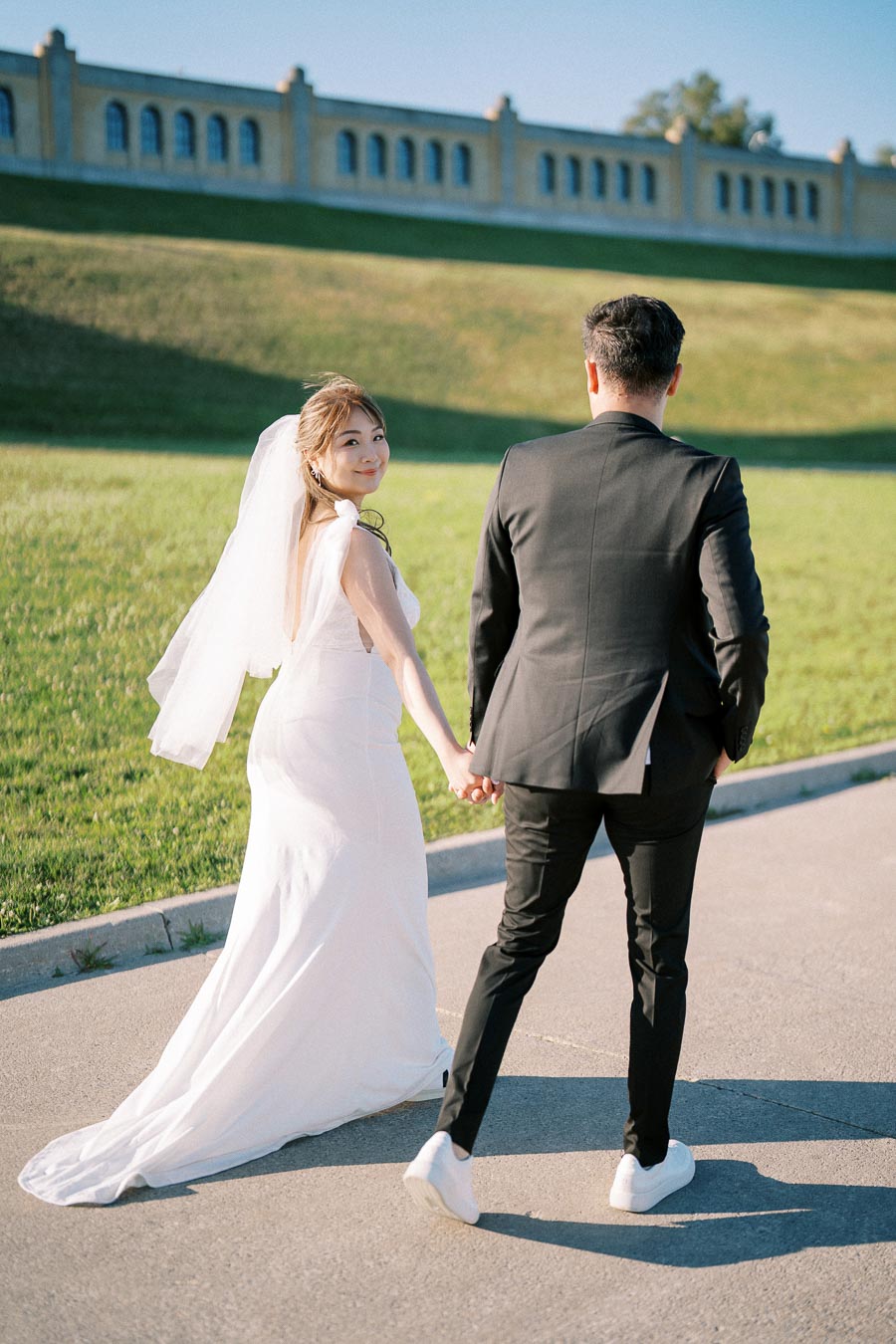 Bride and groom hold hands walking outside, bride in white gown smiling back, set against a sunny landscape with green grass and a building in the background.