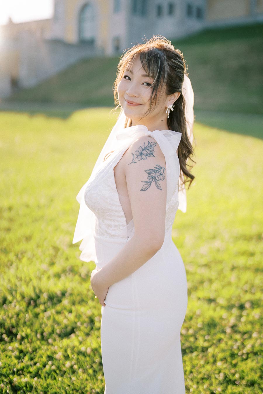 A bride in an elegant white dress poses outdoors, smiling warmly at the camera, with a floral tattoo on her shoulder, against a backdrop of lush green grass and a historic building.