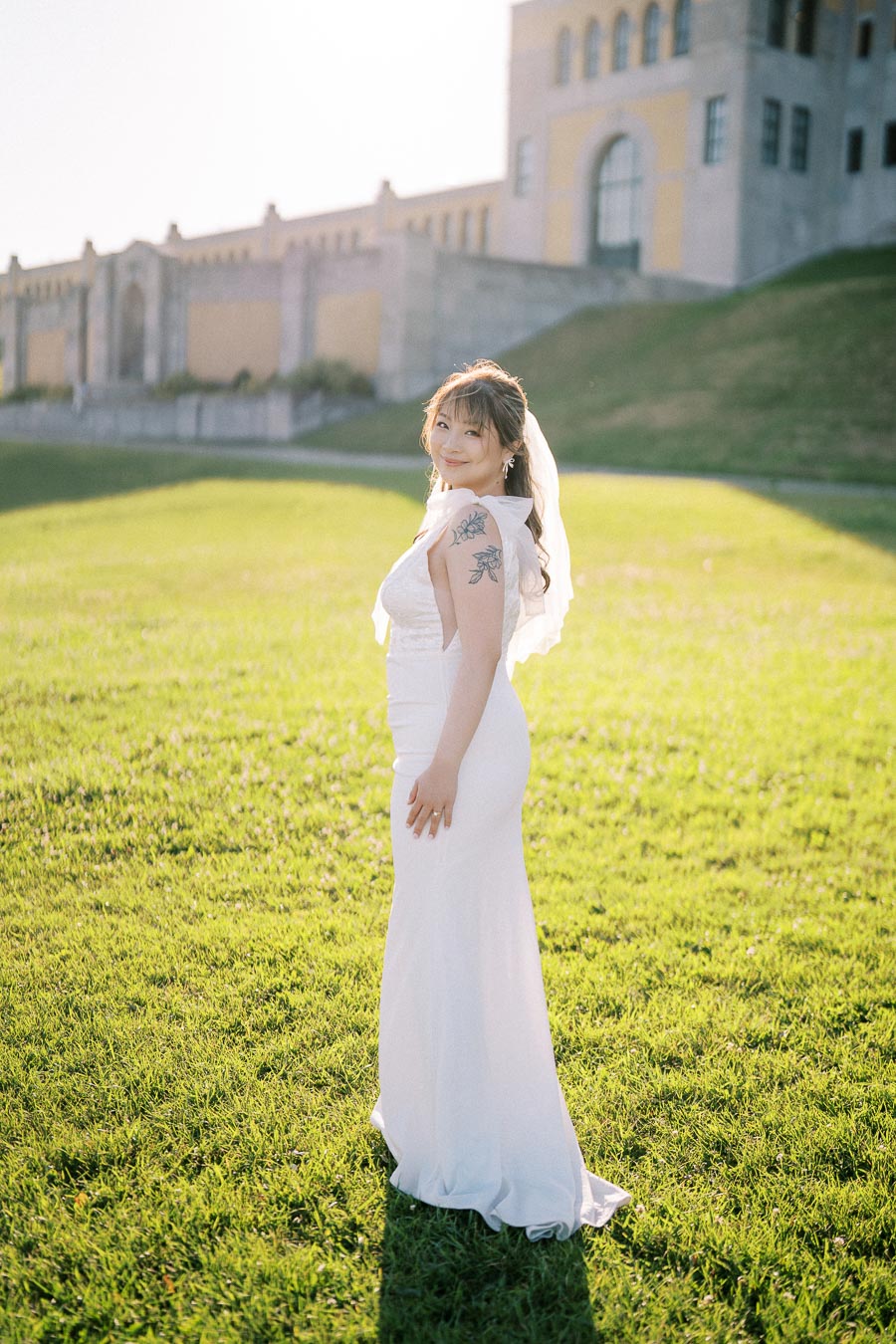 Bride in elegant white wedding dress with floral tattoo, standing on a lush green lawn in front of a historic building, bathed in soft sunlight.