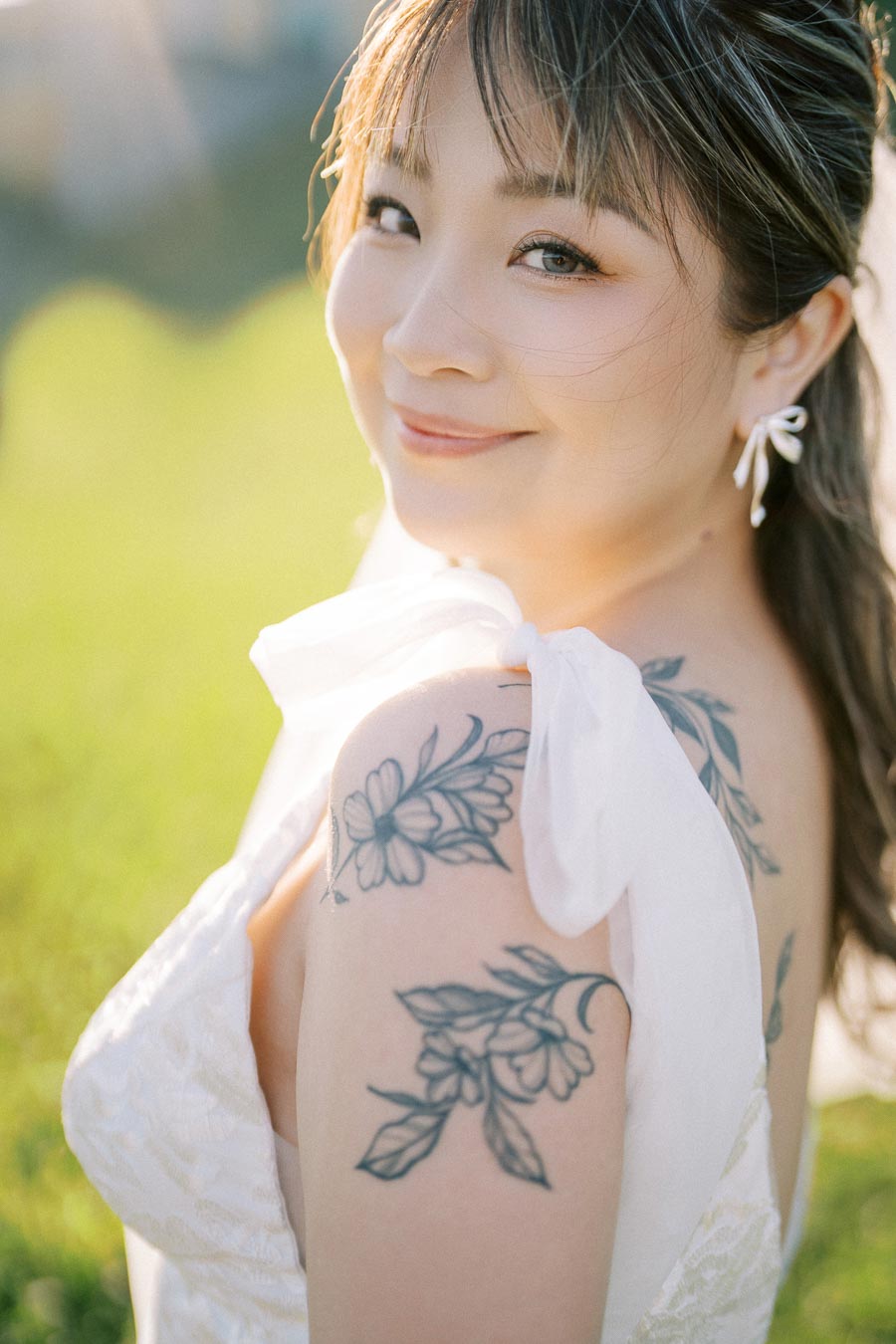 A smiling bride with floral tattoos on her shoulder, wearing a white dress, outdoors in a sunlit setting.