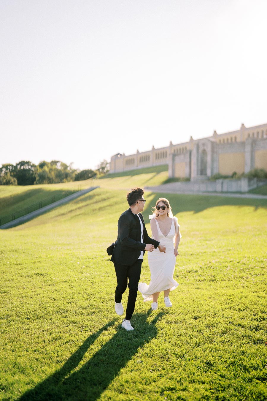 A couple joyfully runs hand-in-hand across a sunlit grassy field beside a historic building, wearing stylish wedding attire.