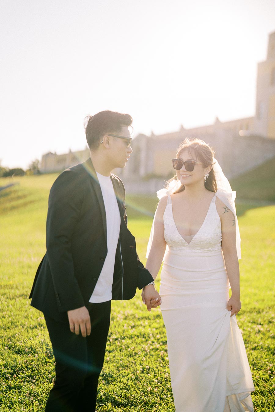 A couple holding hands and smiling in a sunny outdoor setting, with the woman wearing a wedding dress and sunglasses, and the man in a black suit jacket.
