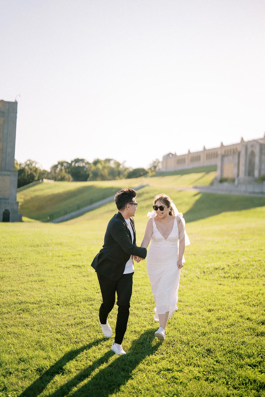 A couple joyfully holding hands and running across a sunlit green field, with elegant historic architecture in the background.