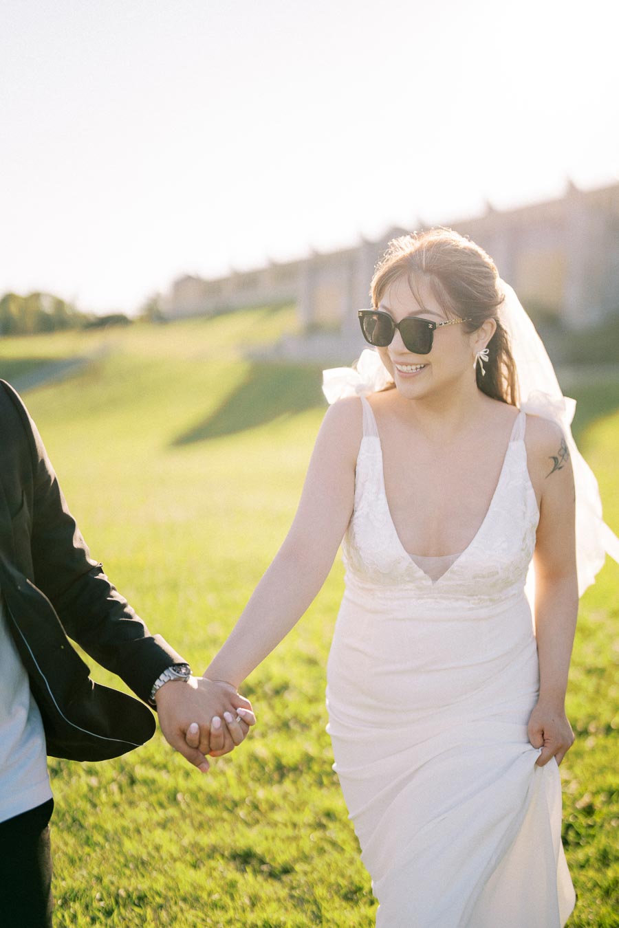 Smiling bride in sunglasses holding hands during outdoor photoshoot in sunlight