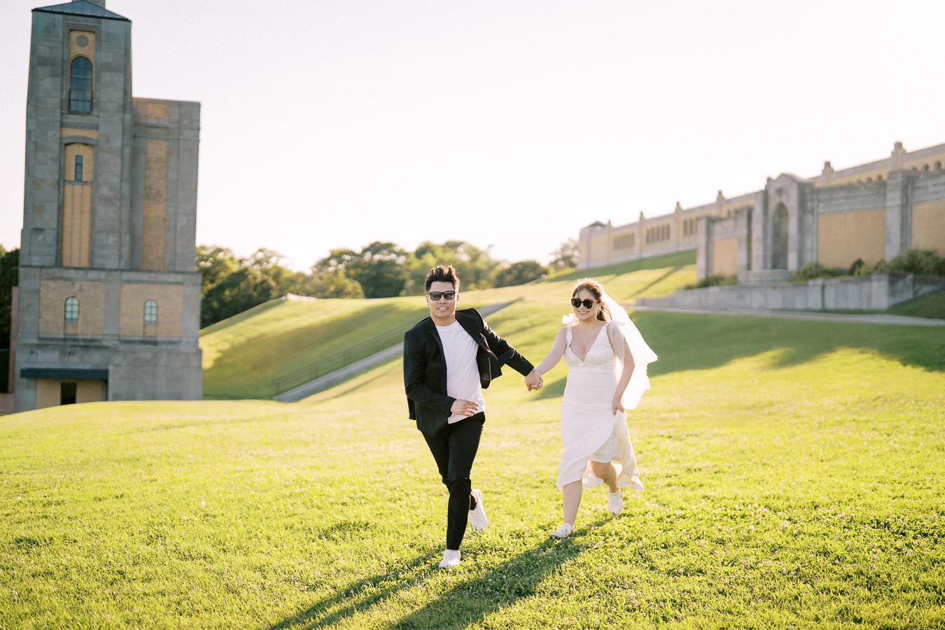 A couple in wedding attire joyfully running across a sunlit grassy field, with a historic building in the background.