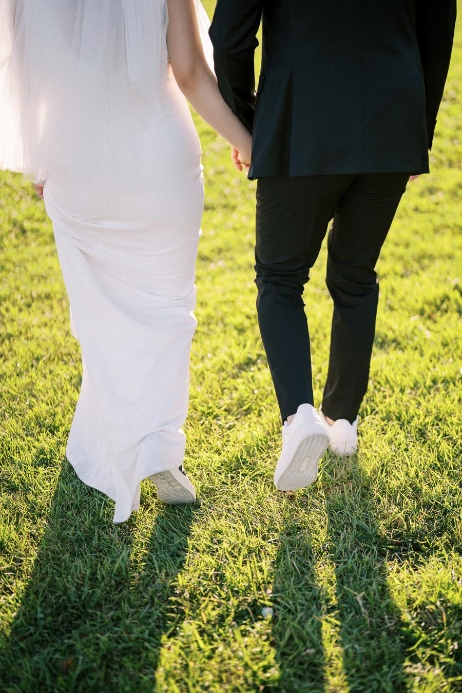 Bride and groom holding hands while walking on grass in wedding attire, captured from behind.
