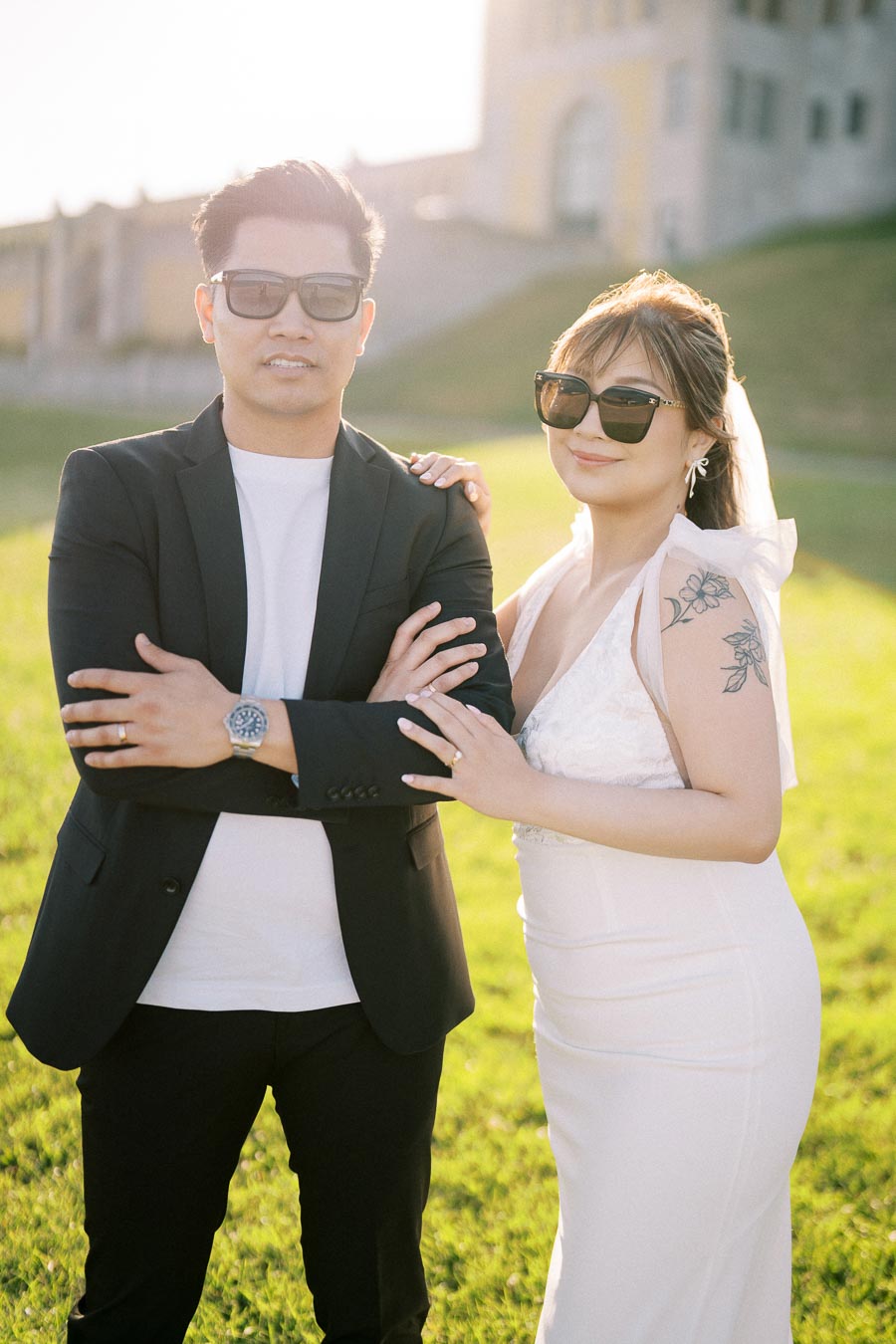 Stylish couple in sunglasses posing outdoors with a blurred castle-like building in the background, woman wearing a white dress with floral tattoo, and man in black blazer and white shirt, enjoying a sunny day in a lush green field.