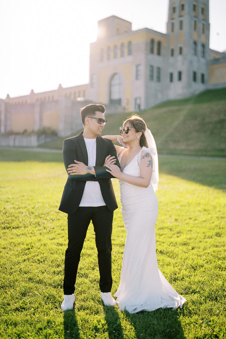 A bride in a white dress and sunglasses stands beside a groom in a black suit and white sneakers, both smiling and enjoying a sunny day on a grassy lawn with an elegant historic building in the background.