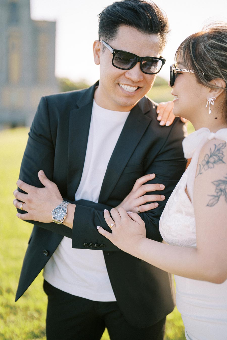 Happy couple wearing sunglasses and formal attire enjoying a sunny outdoor moment, with a focus on their smiles and fashionable accessories.