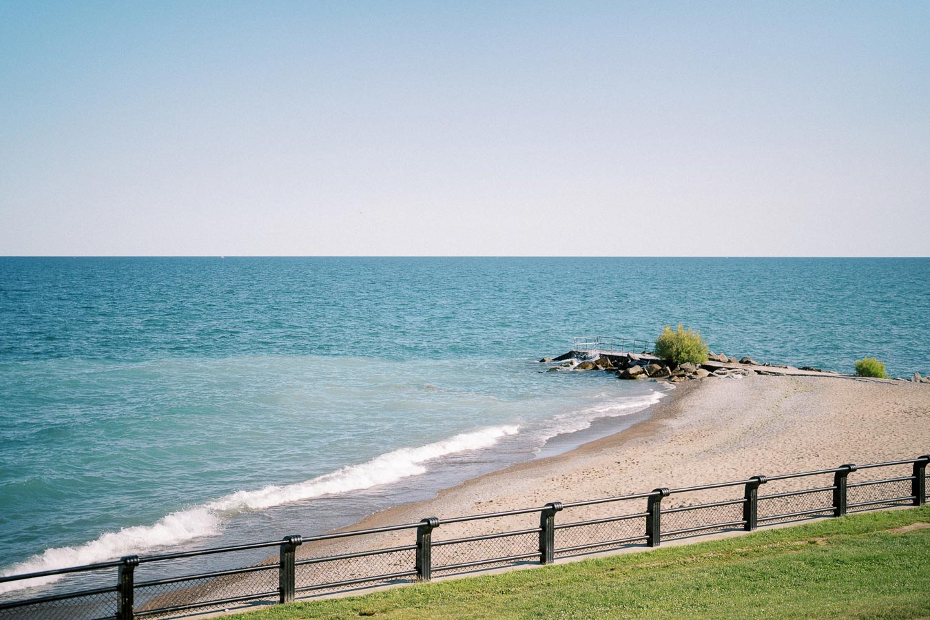 Tranquil coastal scene with blue ocean waves, sandy beach, and a stone jetty bordered by a fence, under a clear sky.