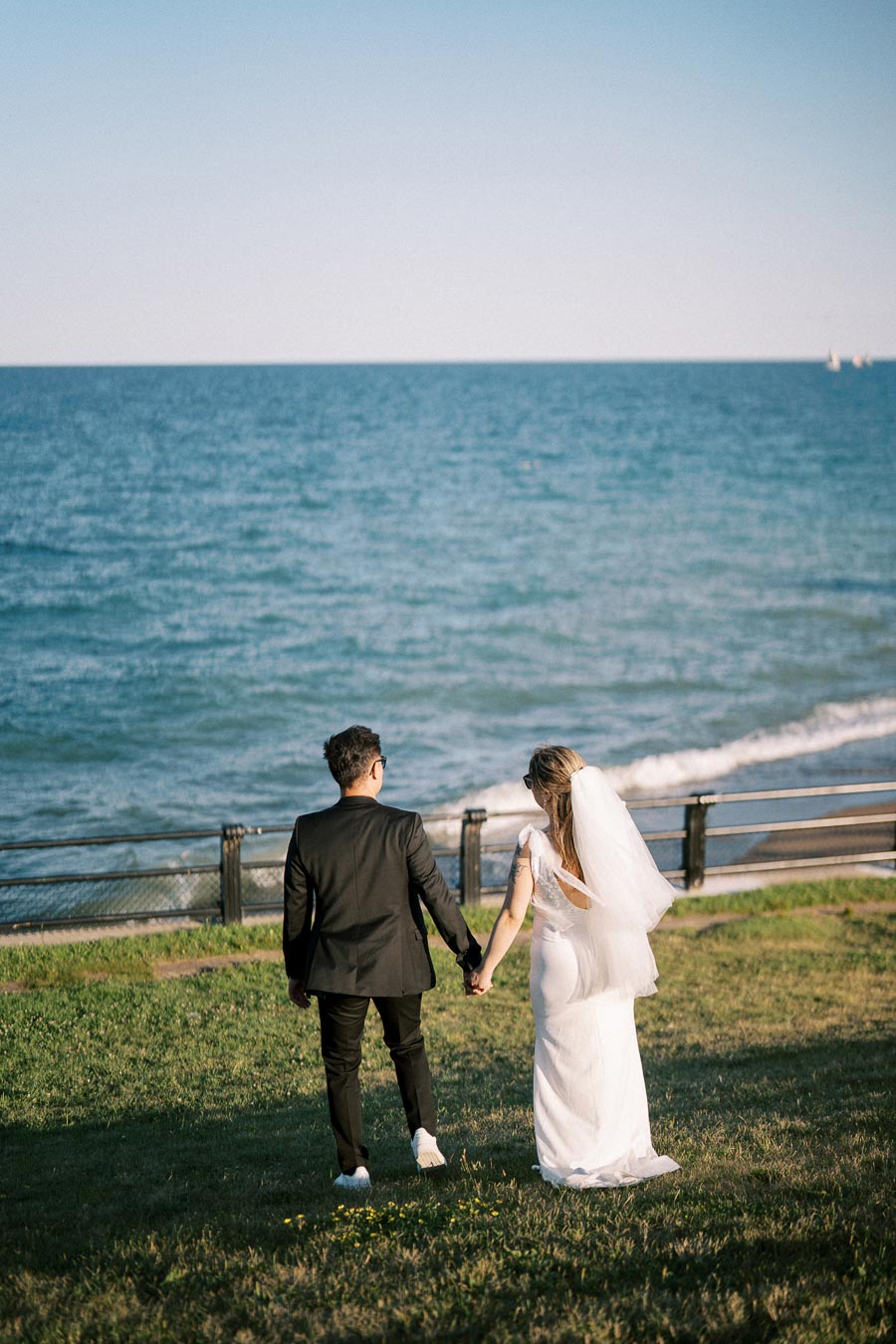 A bride and groom holding hands and walking towards the ocean on a grassy path, with the bride wearing a flowing white gown and veil, and the groom in a black suit, under a clear blue sky.
