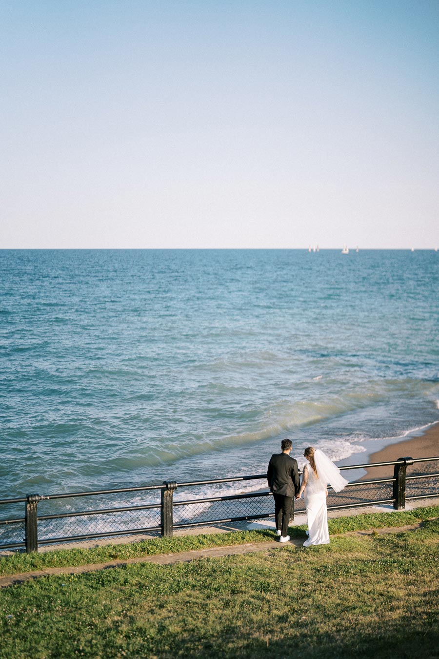Bride and groom walking along a scenic oceanfront path, with a tranquil blue sea and gentle waves in the background.