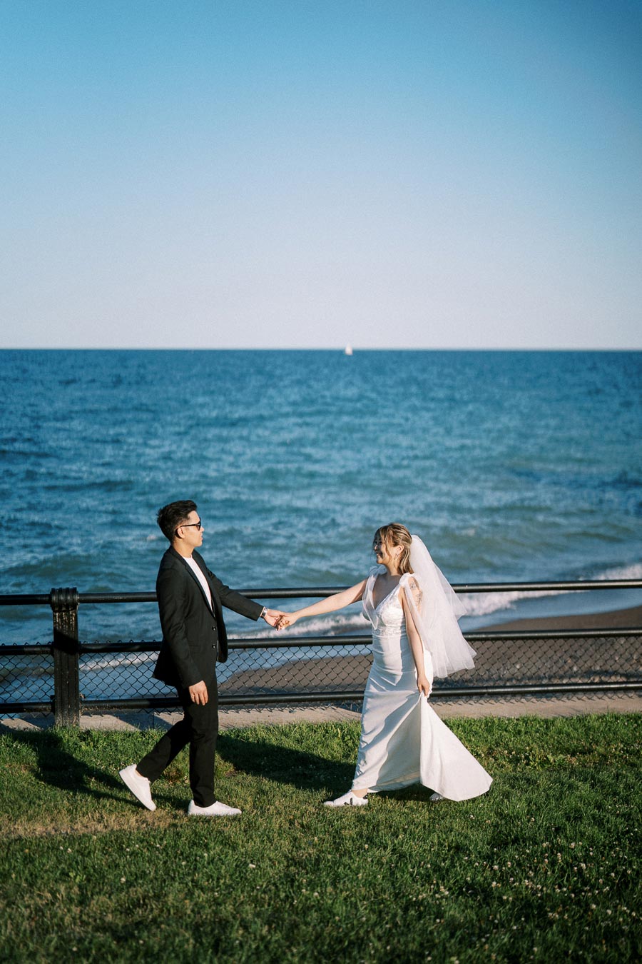 Wedding couple holding hands near the ocean, groom in black suit and bride in white dress with veil, walking by the seaside with clear blue sky.