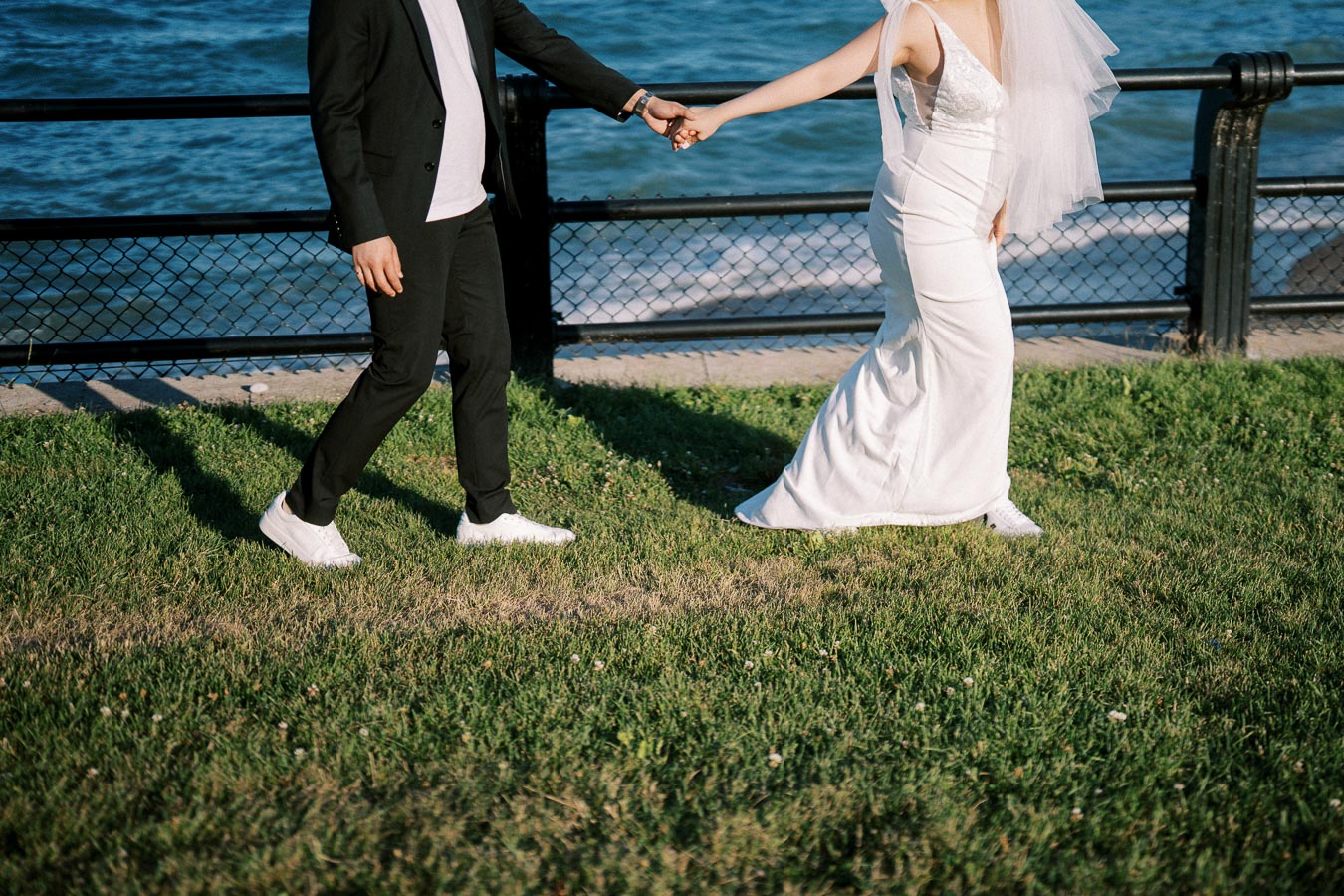 A bride and groom walking hand in hand along a grassy waterfront, with the bride in a white dress and veil, and the groom in a black suit and white sneakers, creating a romantic wedding scene by the water.