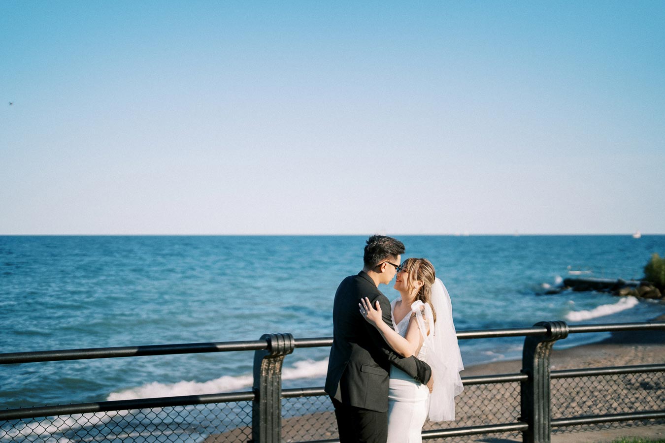 A bride and groom embrace by the ocean on a sunny day, standing near a metal railing with clear blue skies and waves in the background.