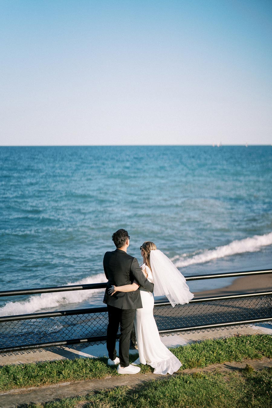 A bride and groom embrace while gazing at the ocean on a sunny day, standing near a metal railing with waves gently crashing below.