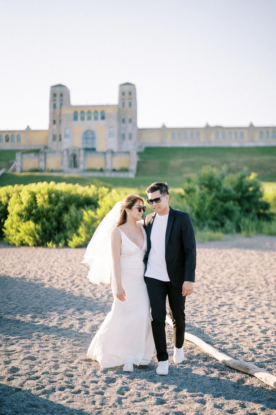 A bride and groom walking on a sunlit beach with a historic building in the background, showcasing a romantic wedding setting.