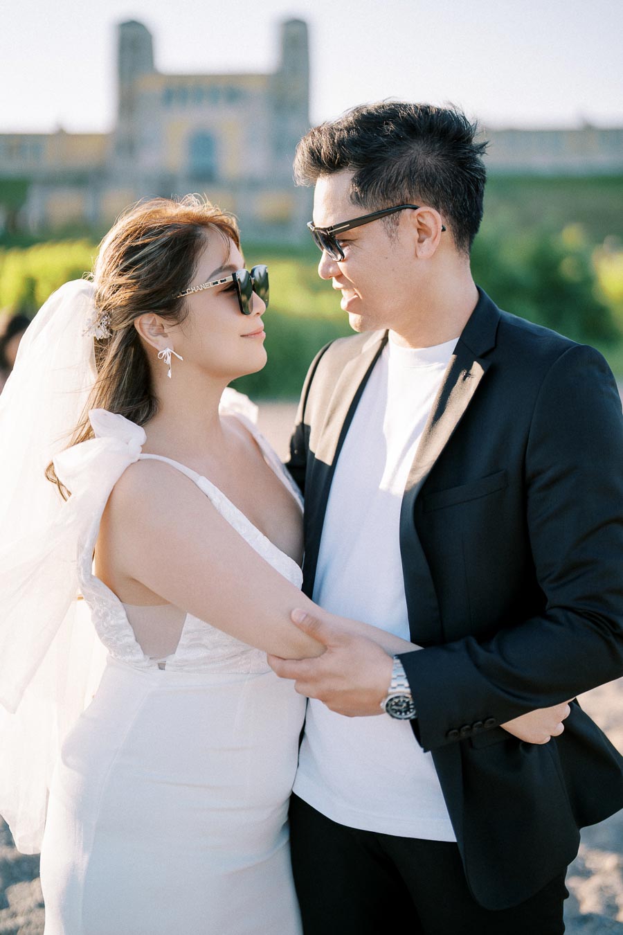 A newlywed couple wearing sunglasses embraces outdoors, with a scenic historical building in the background, creating a romantic wedding moment.