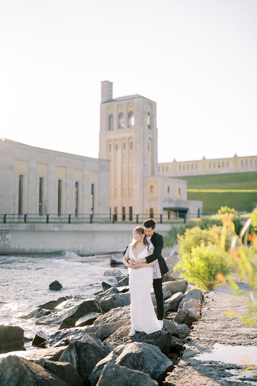 A newlywed couple poses on rocky riverbank in front of a historic industrial building during a sunny day.