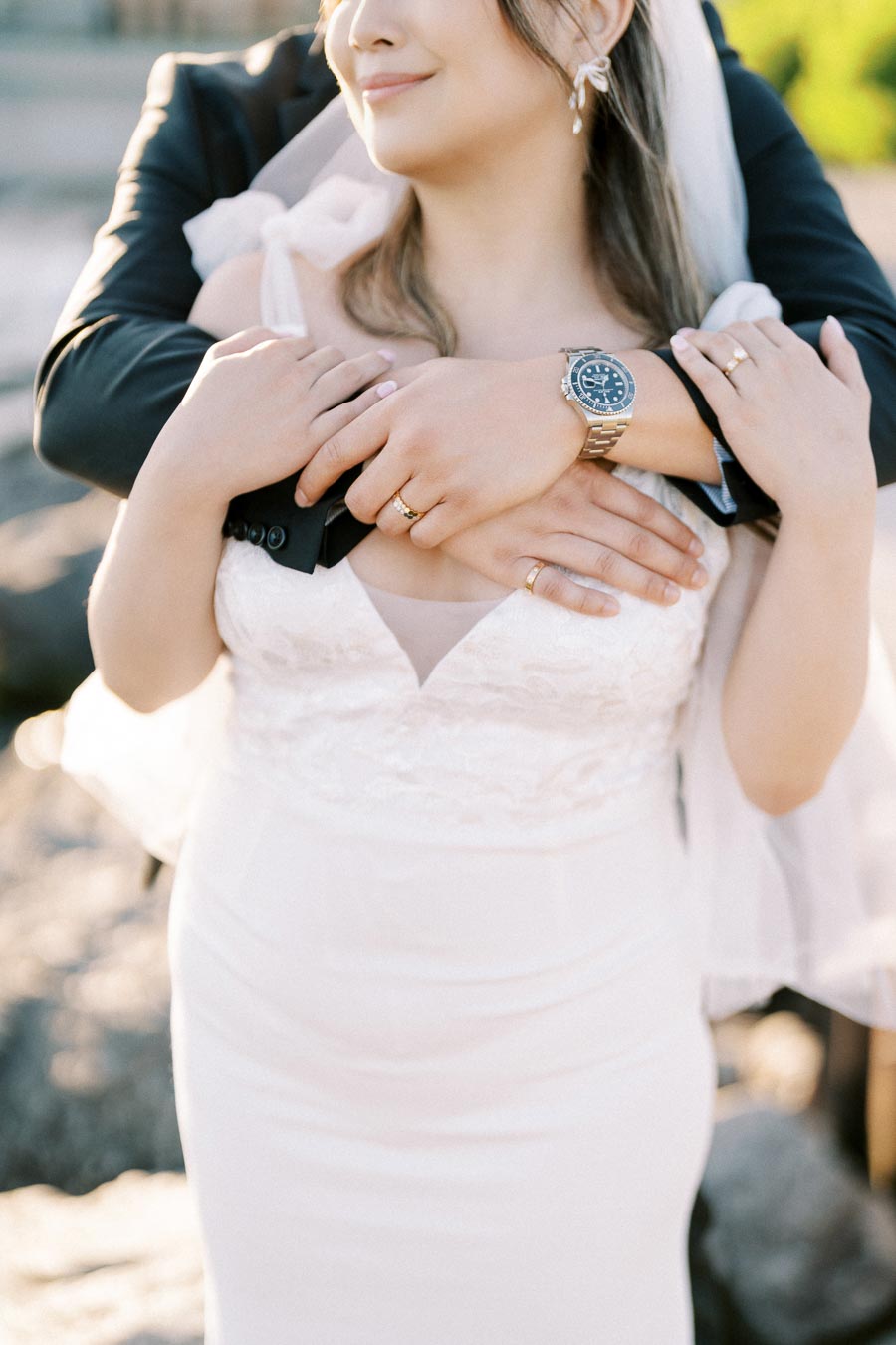 A bride in a white lace wedding dress embraces her partner, who is wearing a watch, during a romantic outdoor photoshoot.
