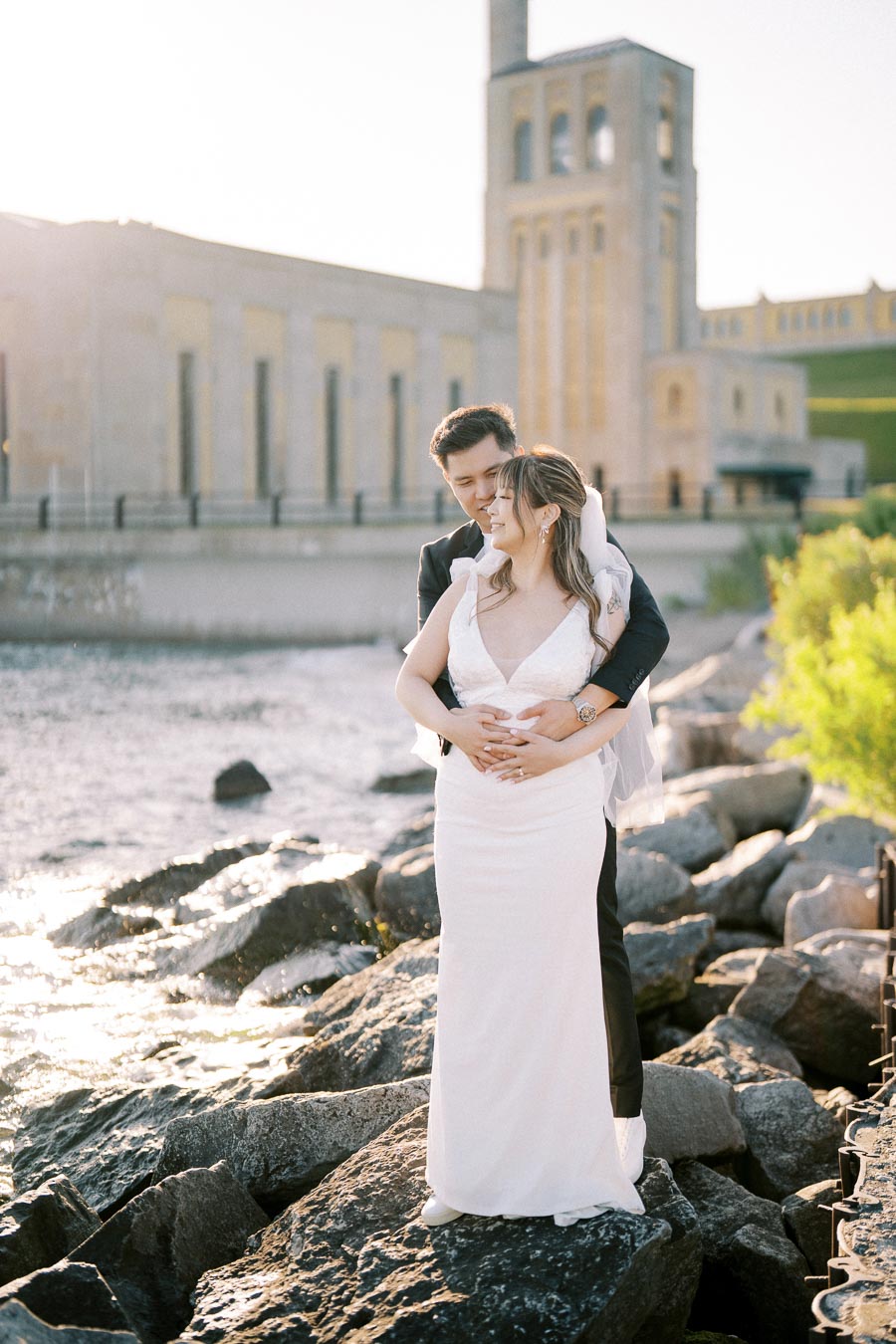 A couple embracing on rocky shore with an industrial building in the background, dressed in formal wedding attire, under bright sunlight.