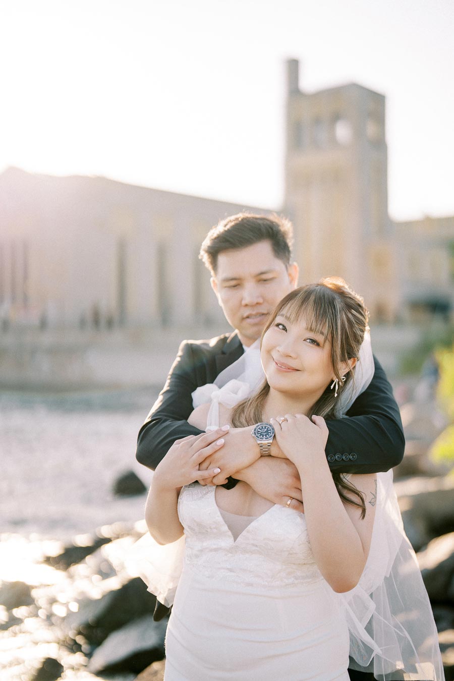 A happy couple embraces by a waterfront, with the bride in a white wedding dress and veil, and the groom in a dark suit, against a sunny backdrop of historic architecture.