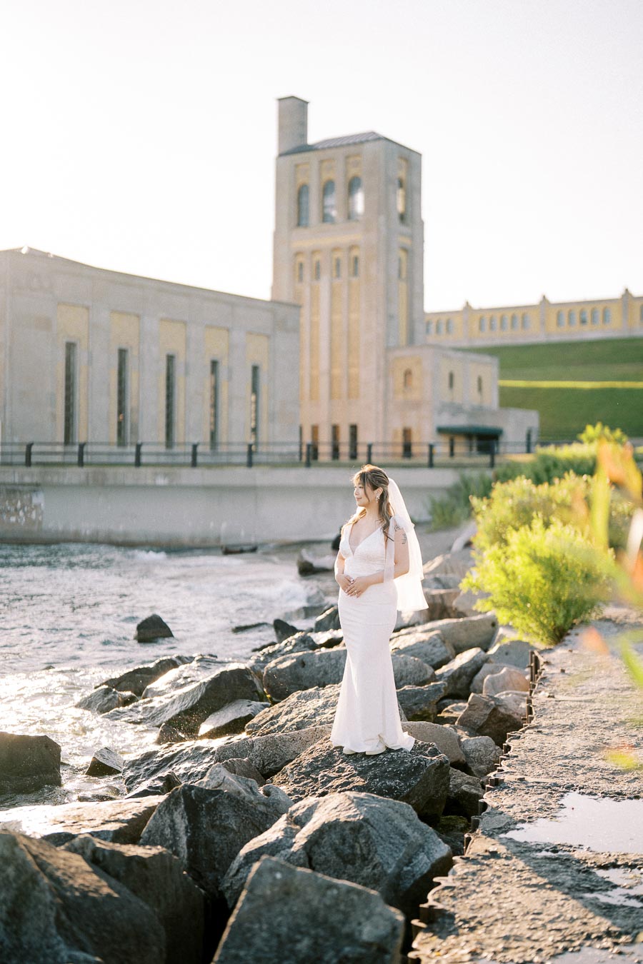Bride in elegant white wedding dress standing on rocky shore with historic building in the background under bright sunlight.