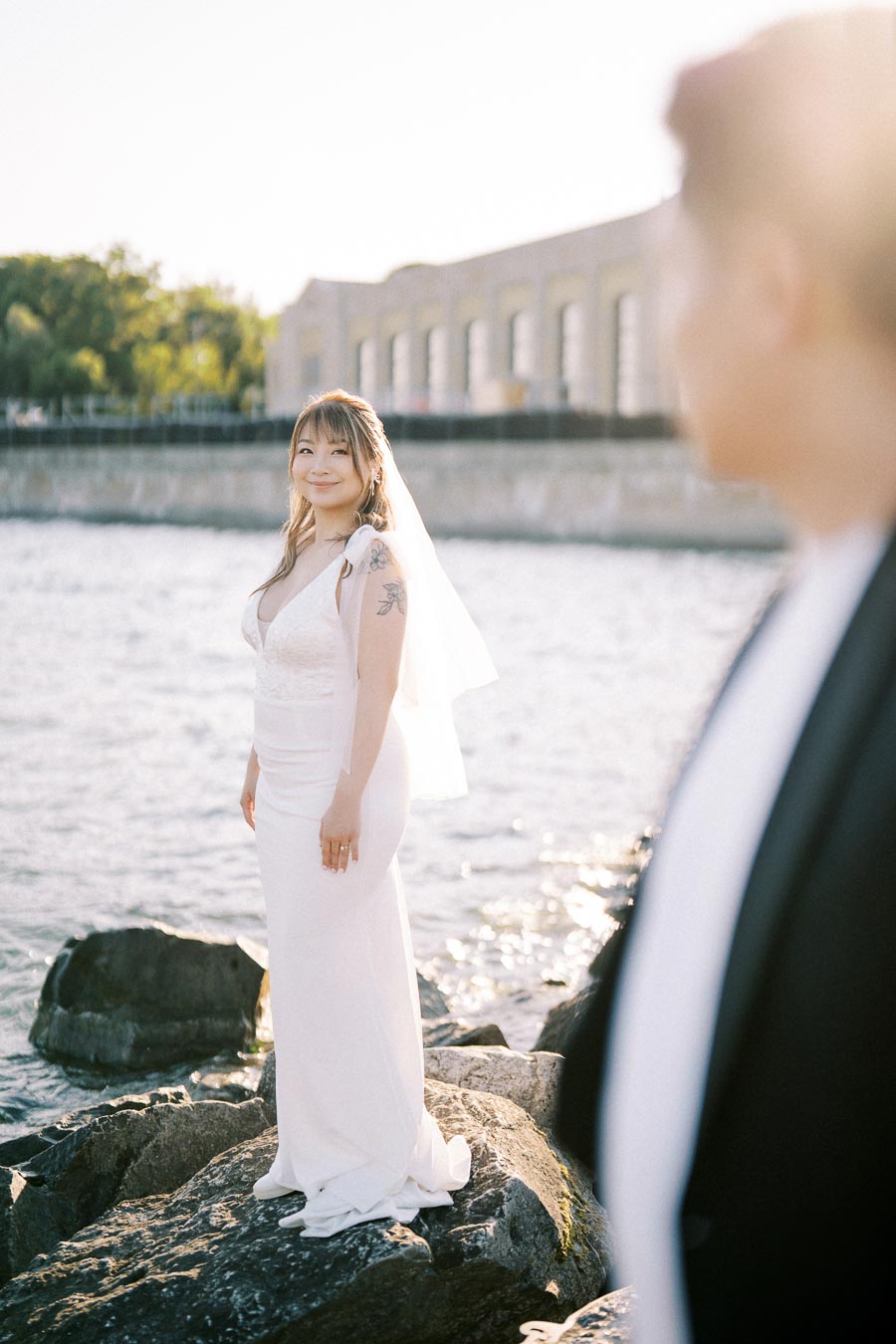 Bride in a white dress standing on rocks by the water, smiling with a scenic waterfront background, featuring soft sunlight and a blurred figure in the foreground.