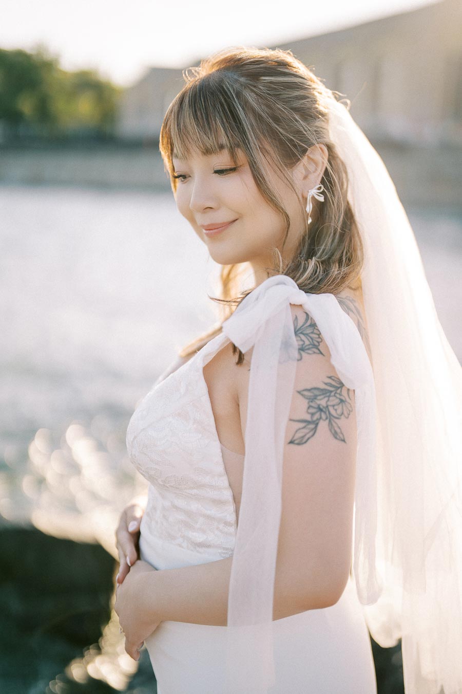 A bride in a white lace wedding dress and veil stands by the water, smiling softly with floral tattoos on her shoulder in a serene outdoor setting.