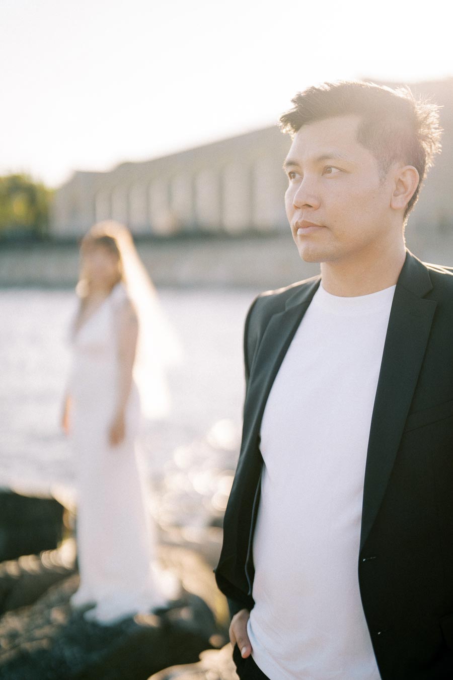 A thoughtful man in a suit standing near a waterfront with a woman in a white dress standing in the background, illuminated by soft sunlight.