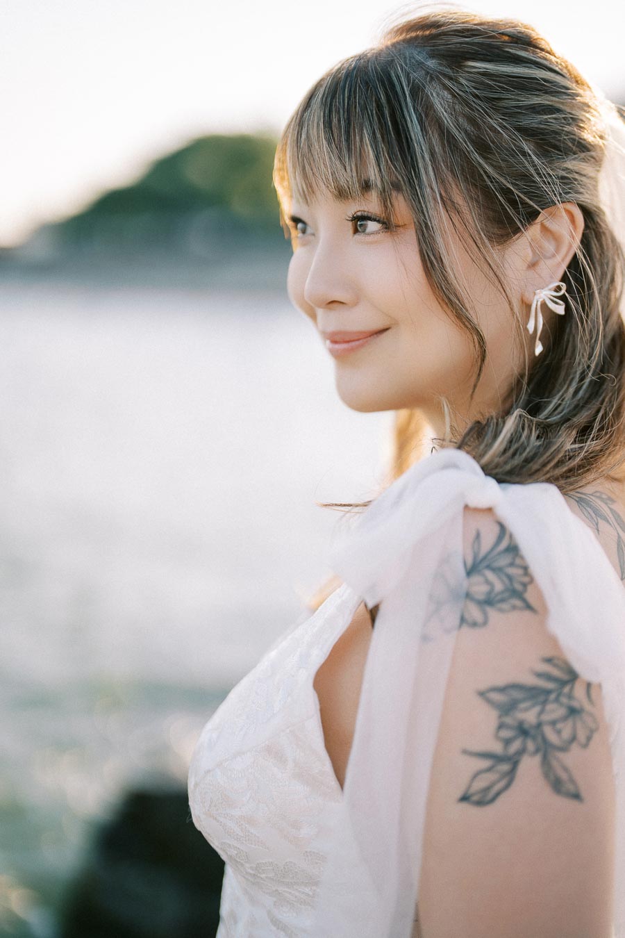 A bride with floral tattoos and pearl earrings gazes serenely into the distance, with an ocean backdrop and soft sunlight highlighting her delicate features.