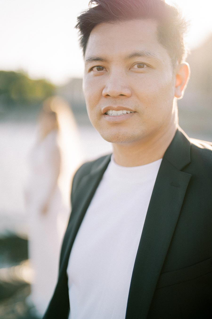 Portrait of a man in a black blazer and white shirt standing outdoors, with a blurred figure in the background and a bright light creating a soft glow.