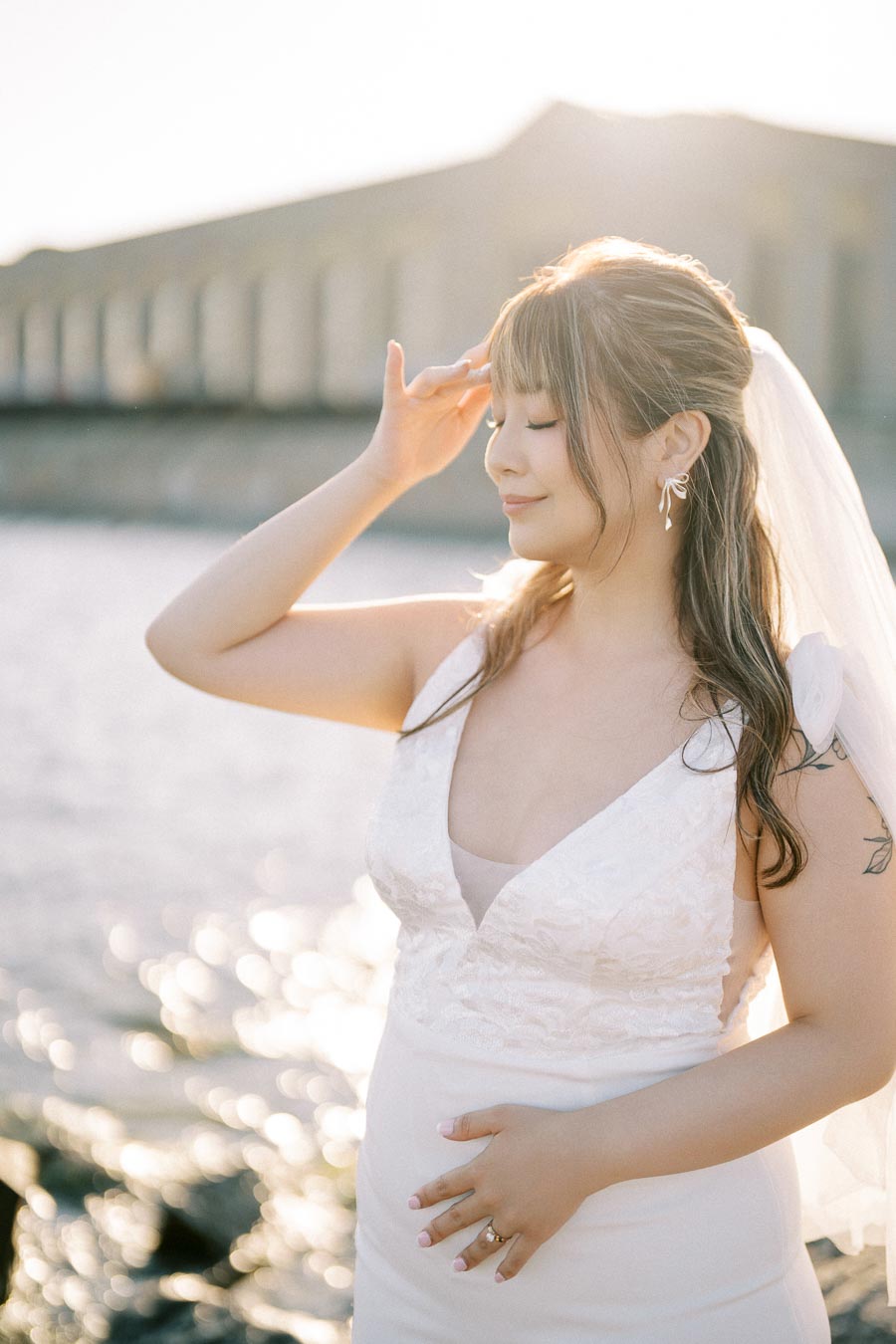 A bride in a white wedding dress posing gracefully by the water, with a sunlit background, wearing a veil and touching her hair gently.